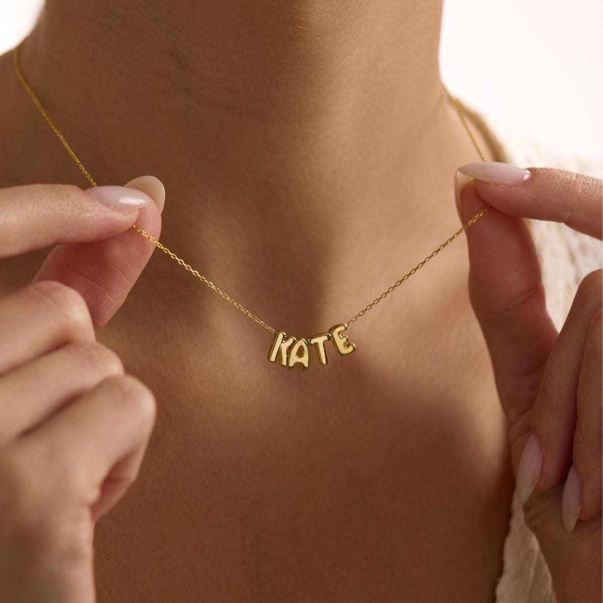 May include: A delicate gold necklace spelling out the name "KATE" in individual block letters. The necklace is held by a person, showcasing the pendant against their skin. The chain is thin and the letters are in a sans-serif font.