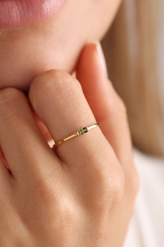 May include: A delicate gold ring featuring a rectangular, olive-green gemstone. The ring's slender band and minimalist design create a refined and elegant aesthetic. The gemstone is approximately 0.25 inches in length.