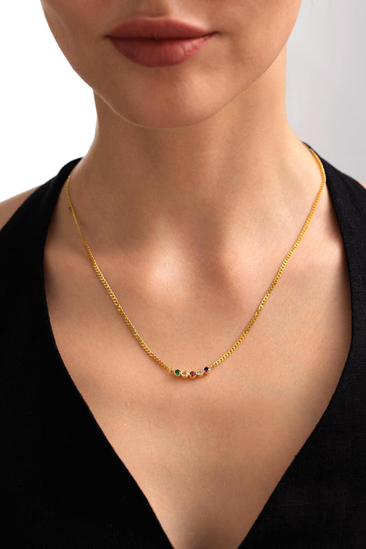 May include: A gold-colored chain necklace with a series of small, colorful gemstones. The gemstones are arranged in a row, with green, orange, and purple stones visible. The necklace is worn against a black top.