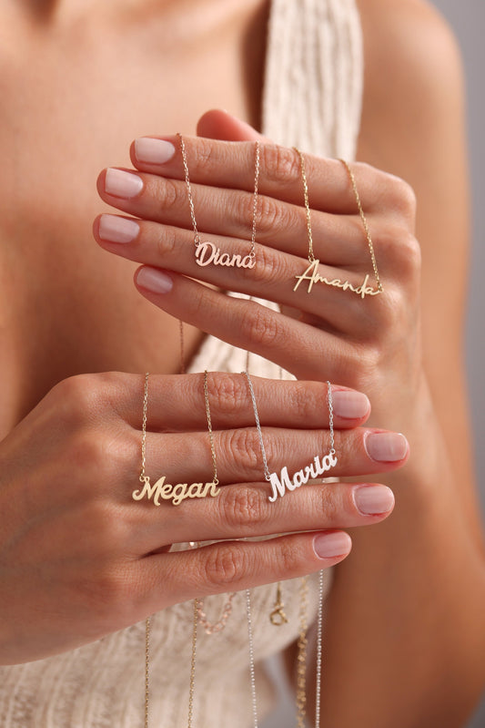 May include: Gold and silver name necklaces with delicate chain. The names "Diana", "Amanda", "Megan", and "Maria" are displayed in a cursive font. The necklaces are held up by a person.