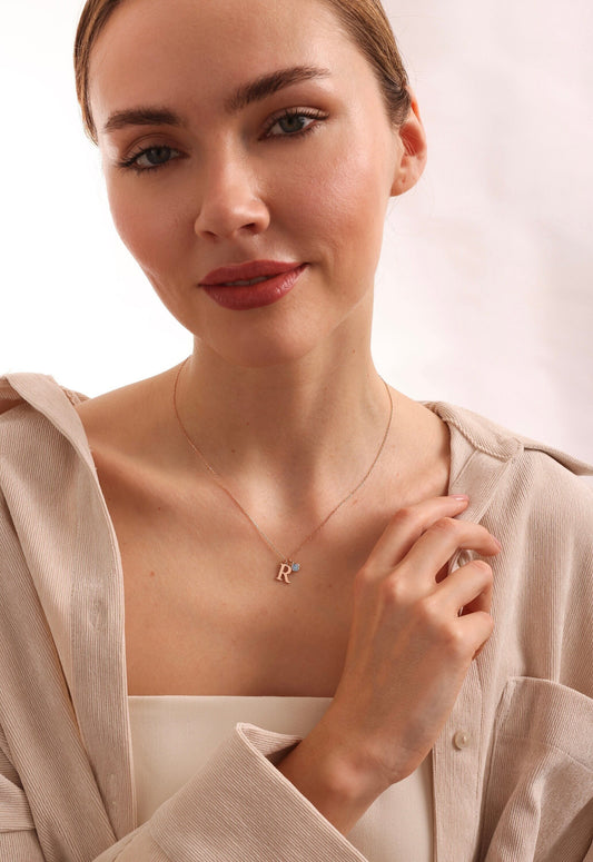 May include: A rose gold necklace with a letter 'R' charm and a small blue gemstone accent.