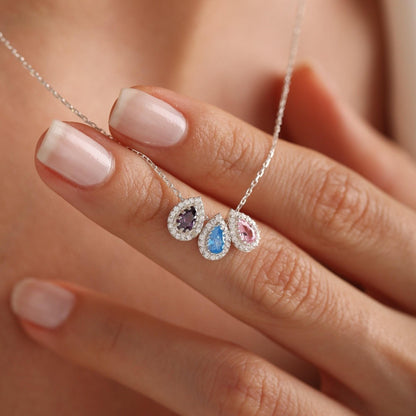 May include: A silver necklace featuring three teardrop-shaped pendants. Each pendant is a different color: purple, blue, and pink, and is surrounded by small, clear stones. The necklace is held by a person.
