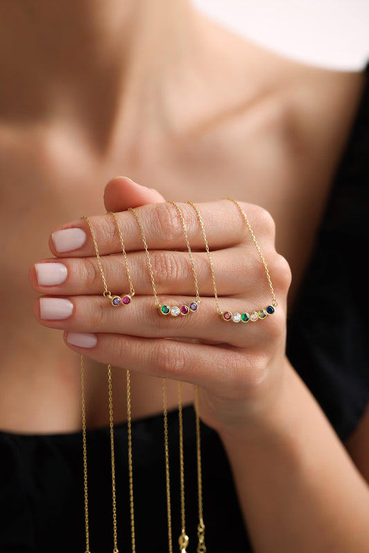 May include: A hand displays several gold necklaces, each featuring a delicate chain and a curved bar pendant. The pendants are adorned with colorful gemstones, including red, purple, green, white, and blue, creating a vibrant and eye-catching design. The necklaces are a jewelry item.