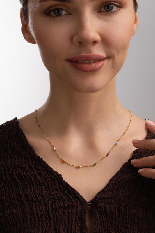 May include: A gold-colored necklace with small, colorful gemstones. The necklace features a delicate chain with a series of tiny, round beads and rectangular gemstones in various colors, including red, green, and blue. The necklace is worn around the neck.