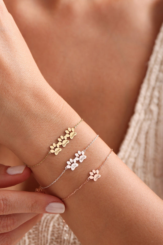 May include: Three gold, silver, and rose gold bracelets with paw print charms. Each charm has a different name engraved on it: Oreo, Scooby, Luna, Lily, Milo, and Lexi.
